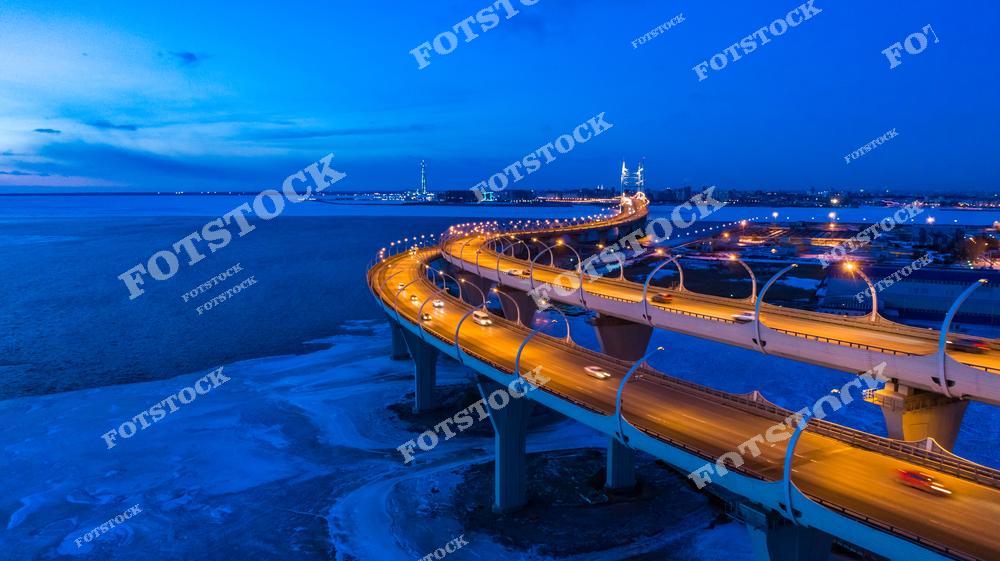 На фотографии изображен участок Западного скоростного диаметра (ЗСД) в Санкт-Петербурге, ведущий вдоль Финского залива. Мост освещен яркими огнями, которые подчеркивают его современные линии. Голубые оттенки ночного неба и залив создают гармоничную и успокаивающую атмосферу.